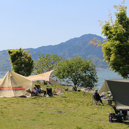 キャンプテントが左右に張られていて右側のテントの前で人が座っている。奥に山が写っており、その手前に川が流れている。